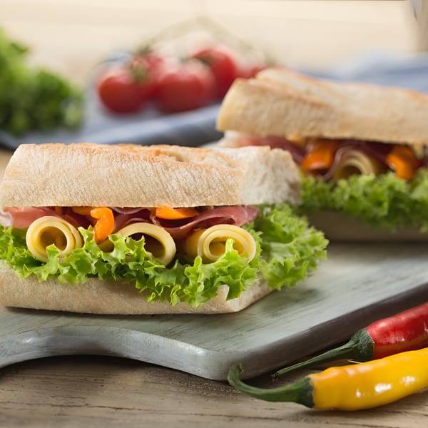 Two ciabatta sandwiches with ham, cheese, lettuce, and peppers on a wooden board.